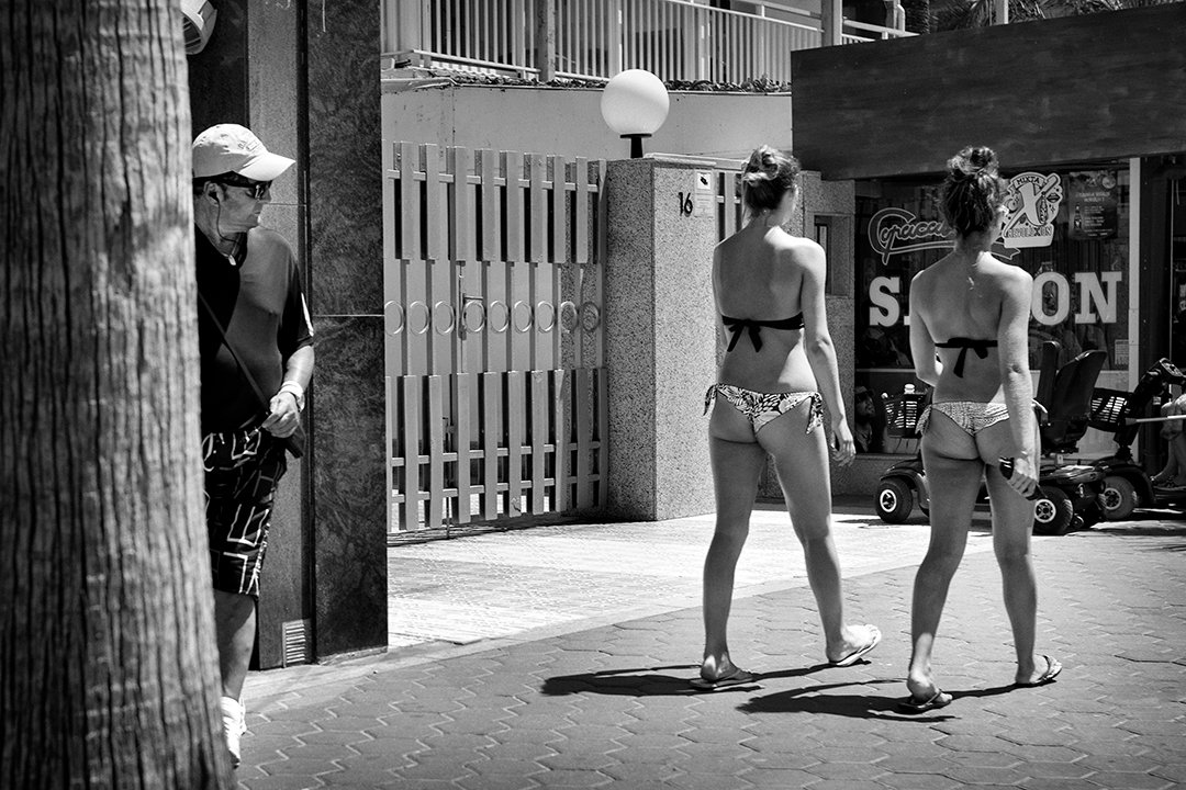 Two people in swimwear beside a palm tree and a facade.